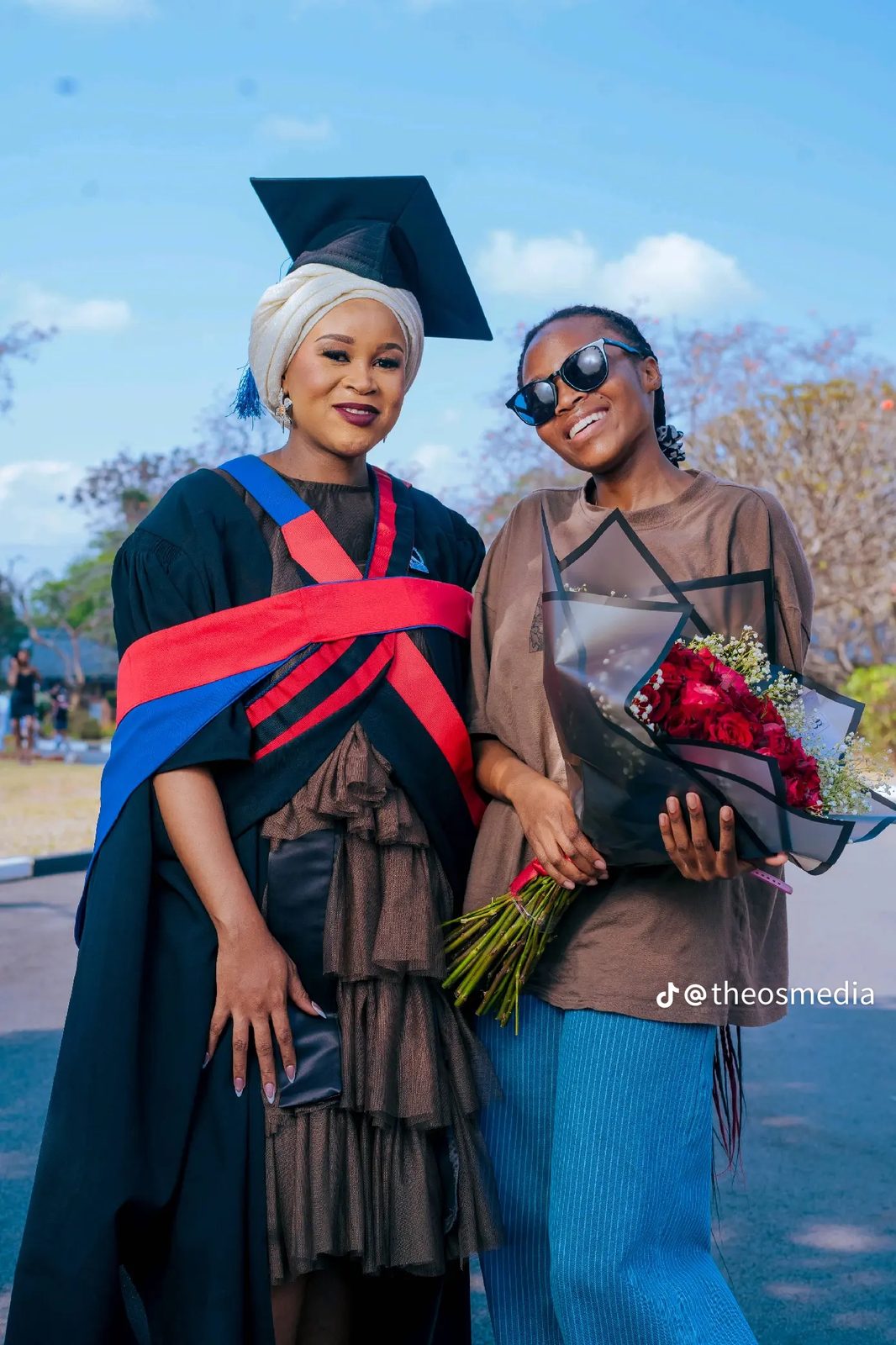 A graduate posing outdoors with a friend holding red roses
