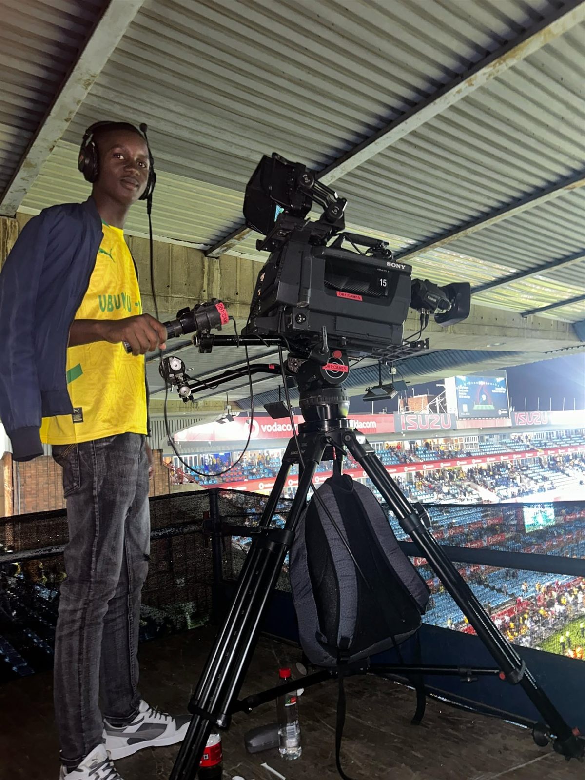 A broadcast camera at a stadium, with a player in a yellow jersey nearby